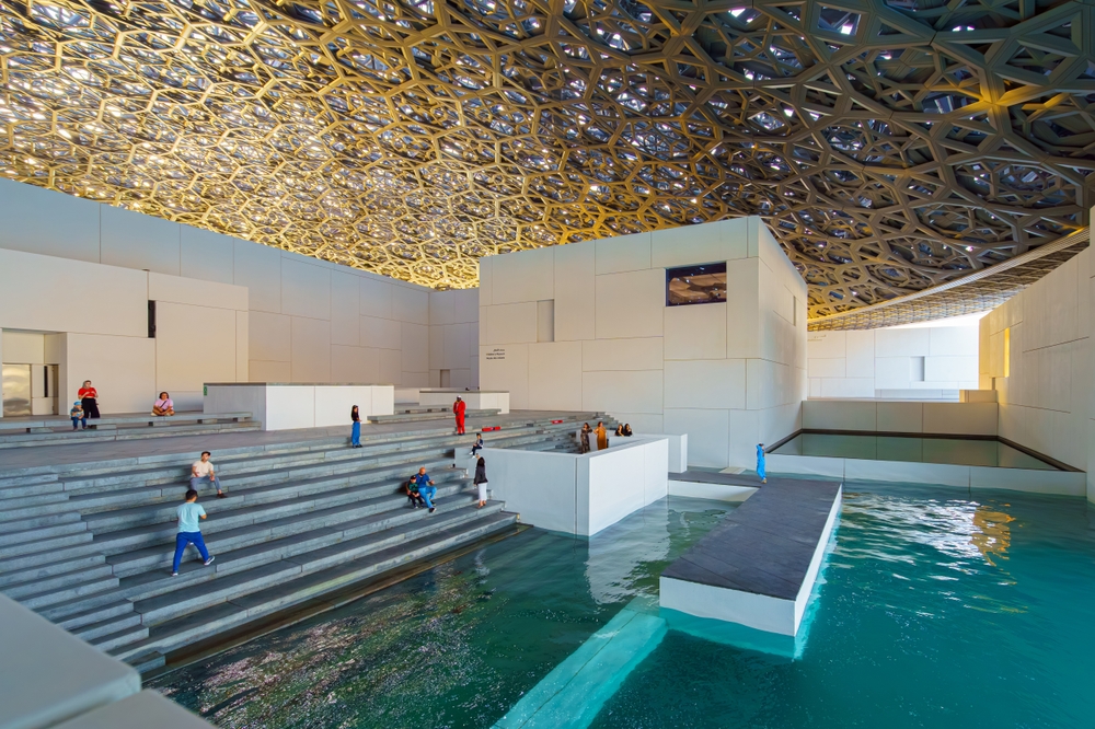 Panoramic view of white concrete buildings and corniche under metal dome of the Louvre Abu Dhabi Museum